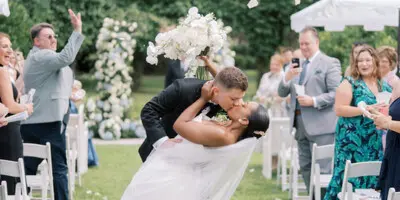 A bride and groom share a kiss during an outdoor wedding ceremony on a grassy lawn, surrounded by guests and white floral arches.