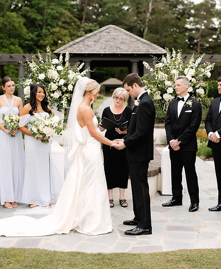 A bride and groom exchanging vows with an officiant during an outdoor wedding ceremony, surrounded by bridesmaids and groomsmen.