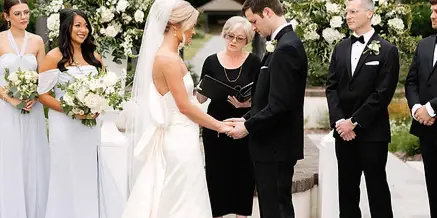 A bride and groom exchanging vows with an officiant during an outdoor wedding ceremony, surrounded by bridesmaids and groomsmen.