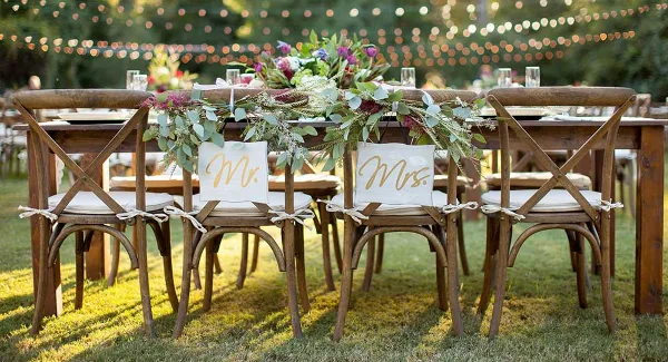 Outdoor wedding head table set with wooden cross-back chairs, white linens, and a floral centerpiece.