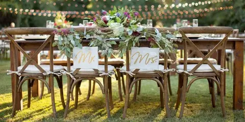Outdoor wedding head table set with wooden cross-back chairs, white linens, and a floral centerpiece.