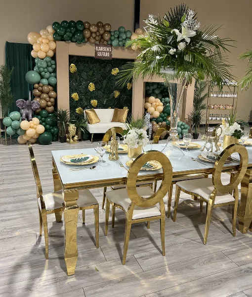 A decorated event venue featuring a gold and white head table, a balloon arch with green and gold balloons, and a jungle-themed backdrop.