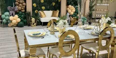 A decorated event venue featuring a gold and white head table, a balloon arch with green and gold balloons, and a jungle-themed backdrop.