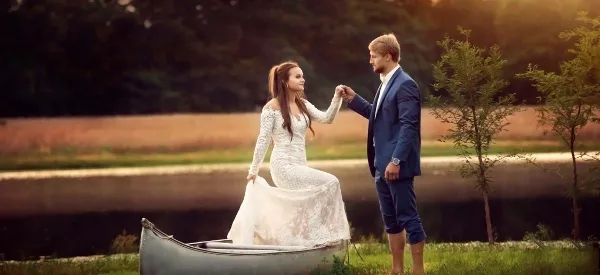 A bride and groom standing by a canoe near a pond in a rural outdoor setting.