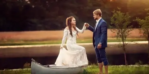 A bride and groom standing by a canoe near a pond in a rural outdoor setting.