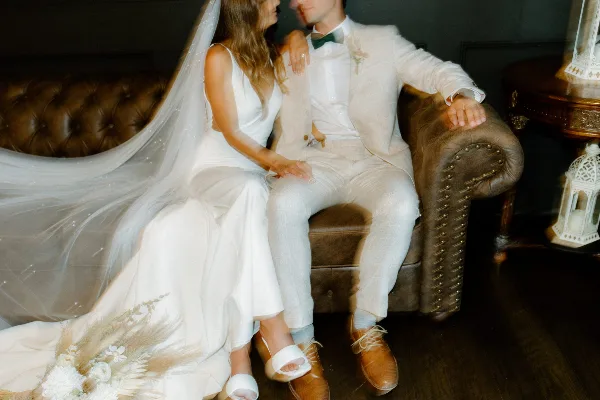 A bride and groom in white wedding attire sitting together on a brown leather sofa.