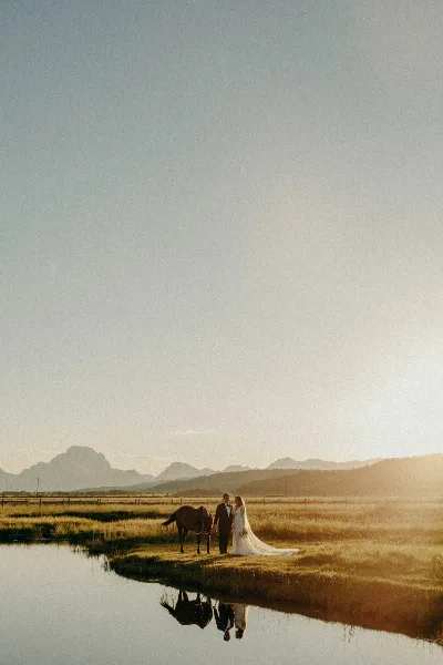 A bride and groom standing with a horse in a grassy field near a body of water with mountains in the background during golden hour.