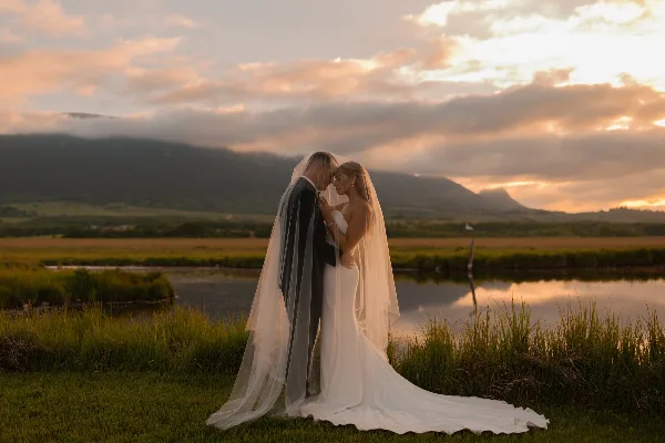 A bride and groom standing close together in a grassy field with a lake and mountains in the background during sunset.