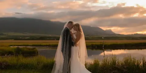 A bride and groom standing close together in a grassy field with a lake and mountains in the background during sunset.