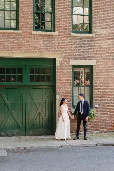 A bride and groom holding hands and smiling while standing in front of a red brick building with green doors.
