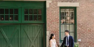 A bride and groom holding hands and smiling while standing in front of a red brick building with green doors.
