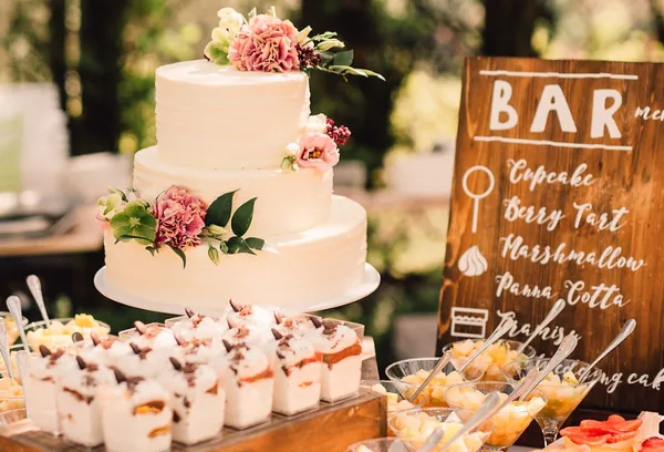 A three-tier white wedding cake decorated with pink flowers sits next to a wooden 'BAR' sign and a display of assorted desserts.