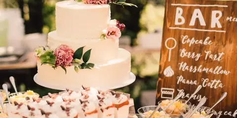 A three-tier white wedding cake decorated with pink flowers sits next to a wooden 'BAR' sign and a display of assorted desserts.