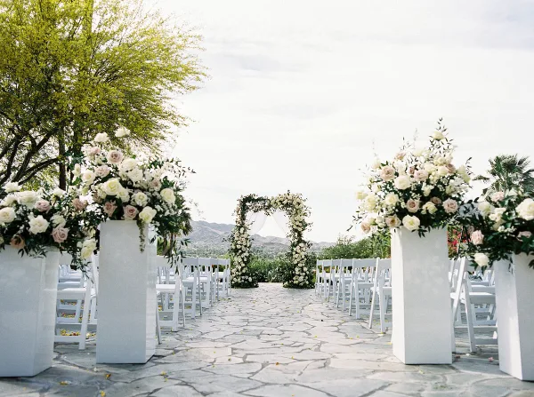 Outdoor wedding ceremony aisle with white chairs, floral pedestals, and a floral arch against a mountain backdrop.