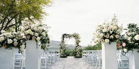 Outdoor wedding ceremony aisle with white chairs, floral pedestals, and a floral arch against a mountain backdrop.