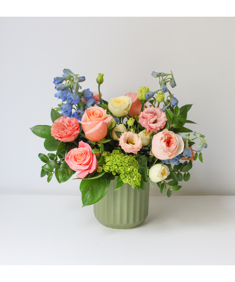 A floral arrangement featuring pink and peach roses, blue delphiniums, and green filler in a light green vase.