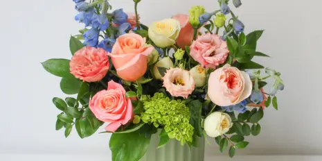 A floral arrangement featuring pink and peach roses, blue delphiniums, and green filler in a light green vase.