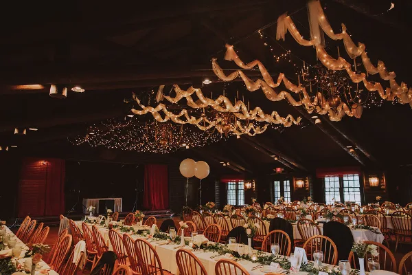 A wedding reception hall featuring long tables with floral centerpieces, wooden chairs, and a ceiling decorated with draped fabric and string lights.