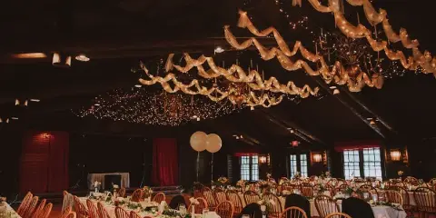 A wedding reception hall featuring long tables with floral centerpieces, wooden chairs, and a ceiling decorated with draped fabric and string lights.