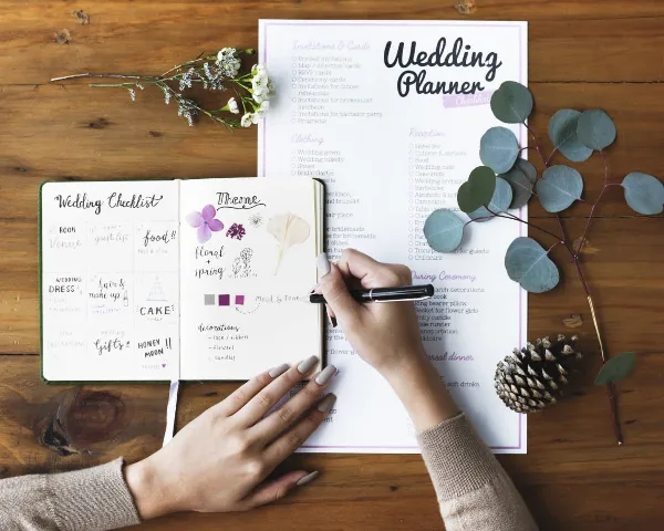 A person's hands writing in a wedding checklist notebook next to a printed wedding planner checklist on a wooden table.