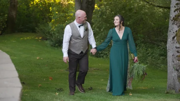A bride in a green dress and a groom in a vest walking hand-in-hand on a grassy path in a wooded area.