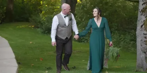 A bride in a green dress and a groom in a vest walking hand-in-hand on a grassy path in a wooded area.