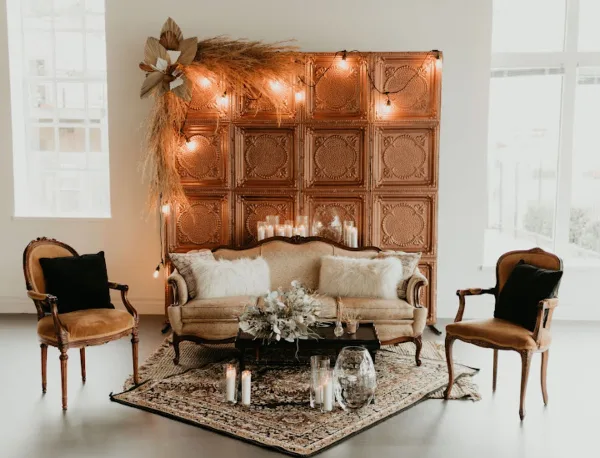 A styled event lounge featuring a vintage sofa, two accent chairs, a patterned rug, and a decorative backdrop with string lights.