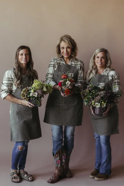 Three women wearing work aprons standing together, each holding a different floral arrangement.