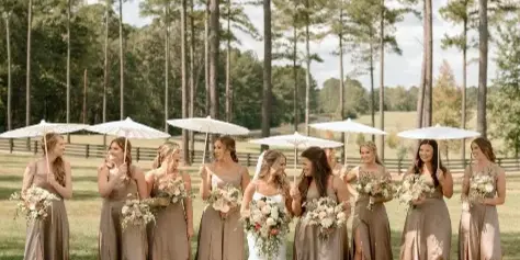 A bride and bridesmaids in taupe dresses holding white parasols and bouquets in a grassy field with a wooden fence and pine trees in the background.