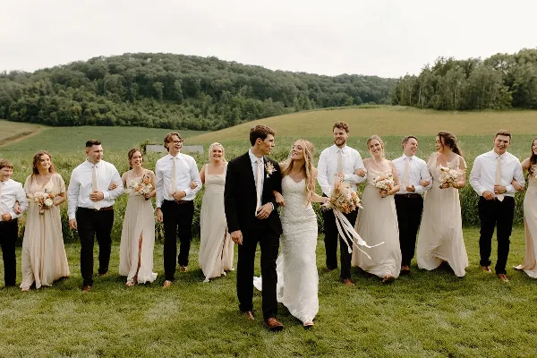 A wedding party including the bride and groom walking through a grassy field with a forested hill in the background.
