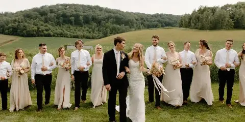 A wedding party including the bride and groom walking through a grassy field with a forested hill in the background.