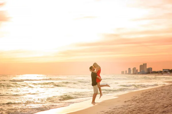 A couple embracing on a sandy beach at sunset with a city skyline in the background.
