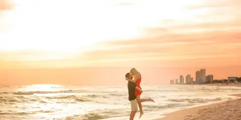 A couple embracing on a sandy beach at sunset with a city skyline in the background.