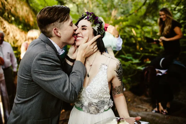 A groom in a grey suit licks cake off the face of a smiling bride wearing a floral crown and white dress in a forest setting.