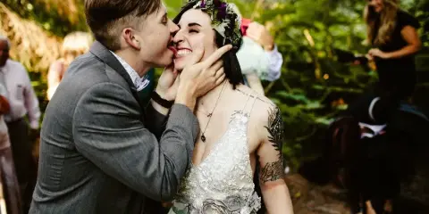 A groom in a grey suit licks cake off the face of a smiling bride wearing a floral crown and white dress in a forest setting.