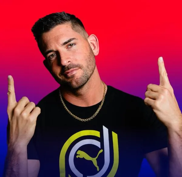 Portrait of a male DJ wearing a black t-shirt with a logo, posing with both index fingers pointing upward against a colorful gradient background.