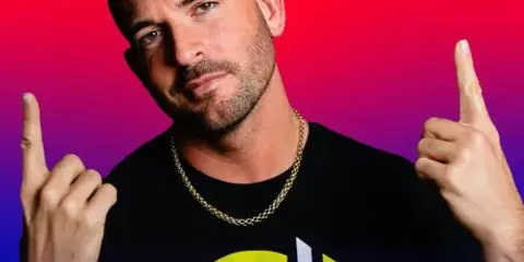 Portrait of a male DJ wearing a black t-shirt with a logo, posing with both index fingers pointing upward against a colorful gradient background.