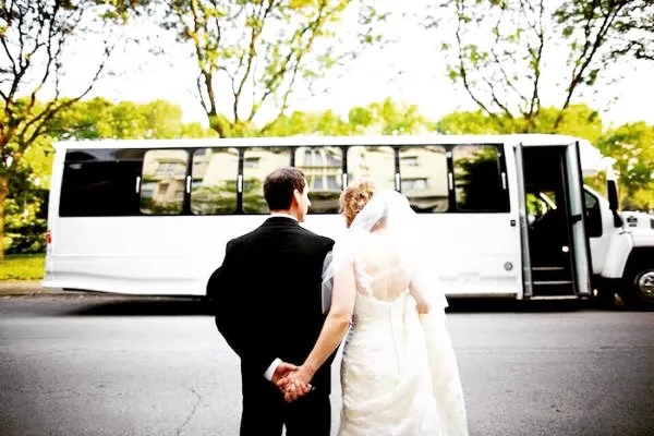 A bride and groom holding hands, viewed from behind, standing in front of a white shuttle bus.