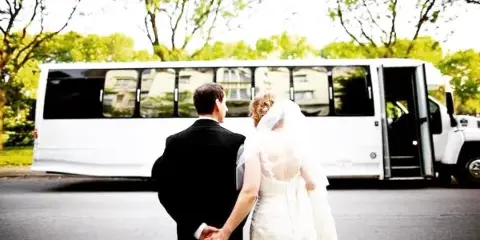 A bride and groom holding hands, viewed from behind, standing in front of a white shuttle bus.