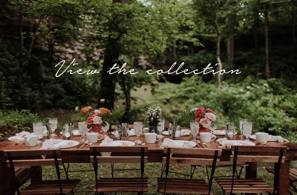 A rustic wooden table set with vintage-style dinnerware, glassware, and floral centerpieces in an outdoor garden setting.