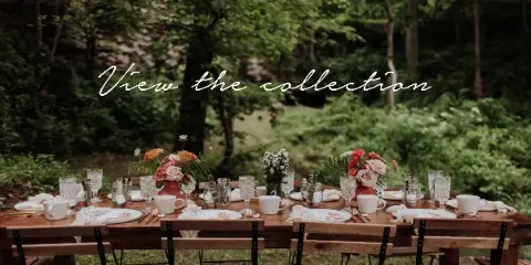 A rustic wooden table set with vintage-style dinnerware, glassware, and floral centerpieces in an outdoor garden setting.