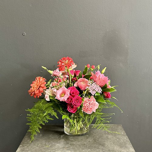 A vibrant floral arrangement with pink and red roses, lilies, and ferns in a clear glass vase against a grey wall.