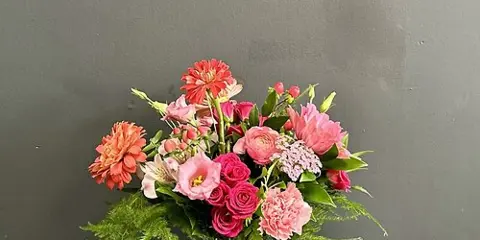 A vibrant floral arrangement with pink and red roses, lilies, and ferns in a clear glass vase against a grey wall.