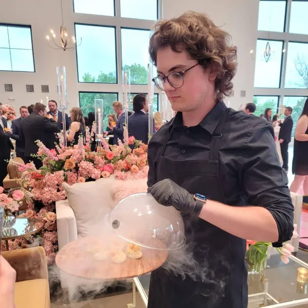 A catering staff member in a black apron and gloves lifts a clear dome from a wooden tray of food at a formal event with floral arrangements and seated guests.