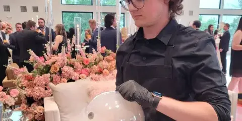 A catering staff member in a black apron and gloves lifts a clear dome from a wooden tray of food at a formal event with floral arrangements and seated guests.