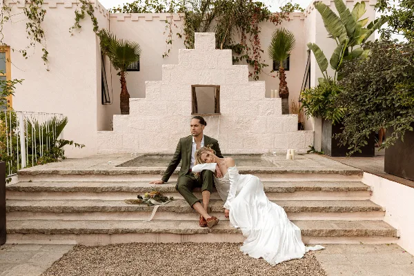 A bride and groom sitting on stone steps in a white-walled courtyard with tropical plants.