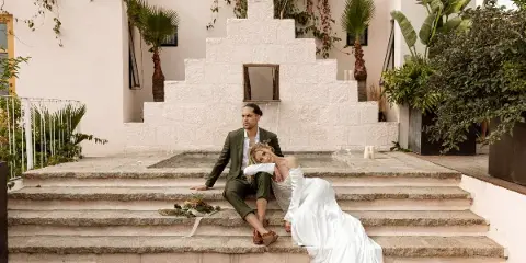 A bride and groom sitting on stone steps in a white-walled courtyard with tropical plants.