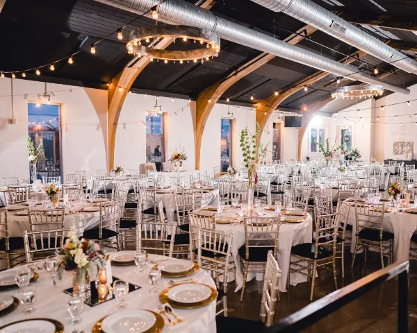Interior view of a wedding reception venue with round tables, white linens, and arched wooden ceiling beams.