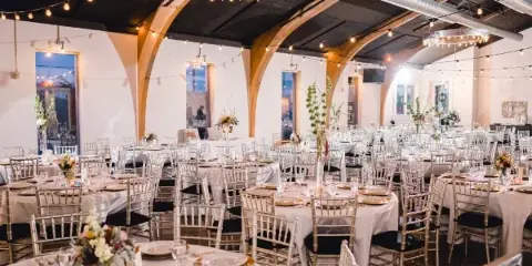 Interior view of a wedding reception venue with round tables, white linens, and arched wooden ceiling beams.