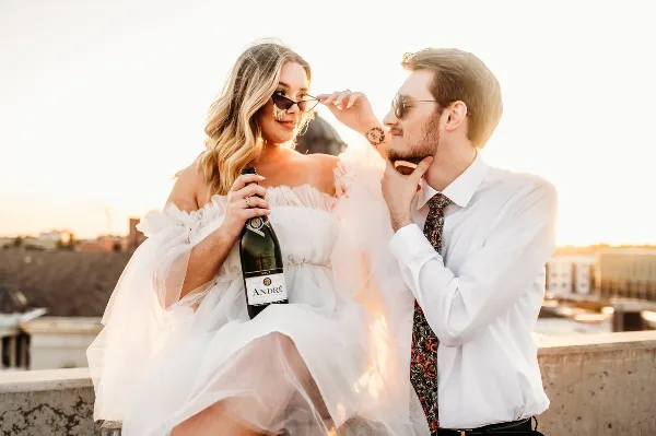 A bride in a white off-the-shoulder gown and a groom in a white shirt and floral tie pose together on a rooftop at sunset, with the bride holding a champagne bottle.